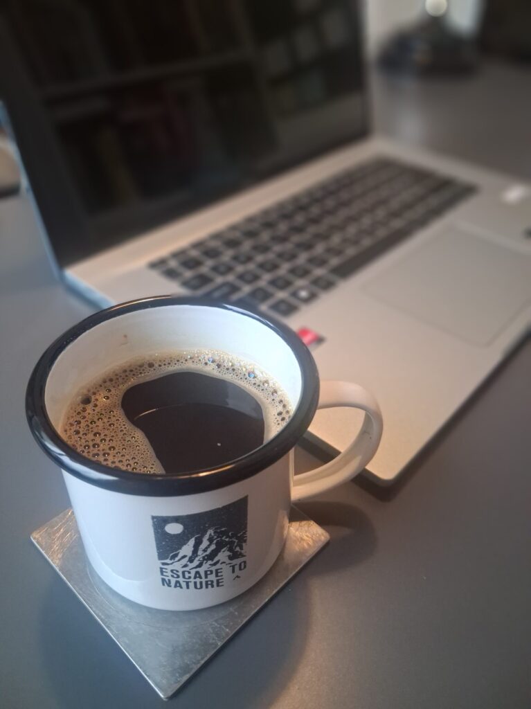 Black coffee in a white enamel mug on a desk next to a laptop in a quiet workspace.